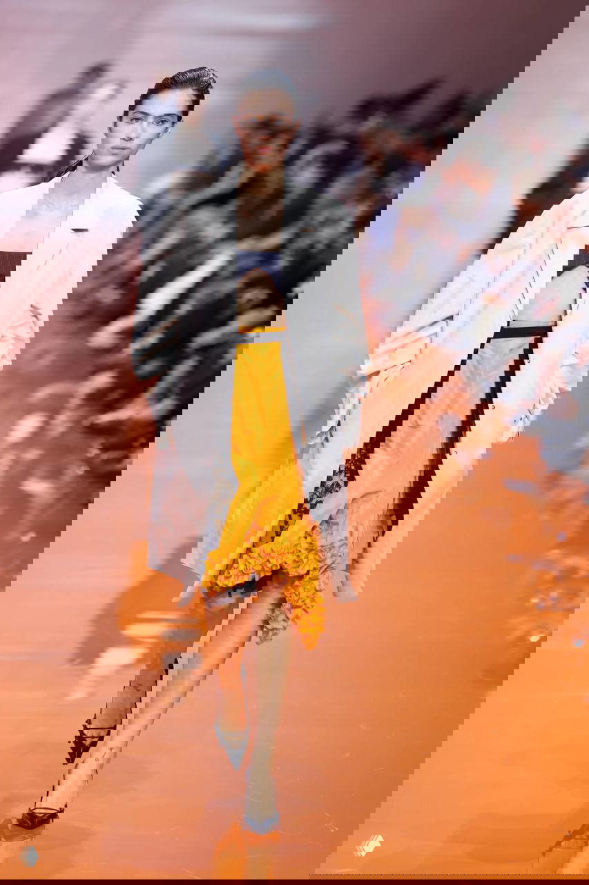 Модель идет по подиуму в белом тренче и яркой желтой юбке с воланами; отражение на глянцевой дорожке усиливает цветовой контраст.