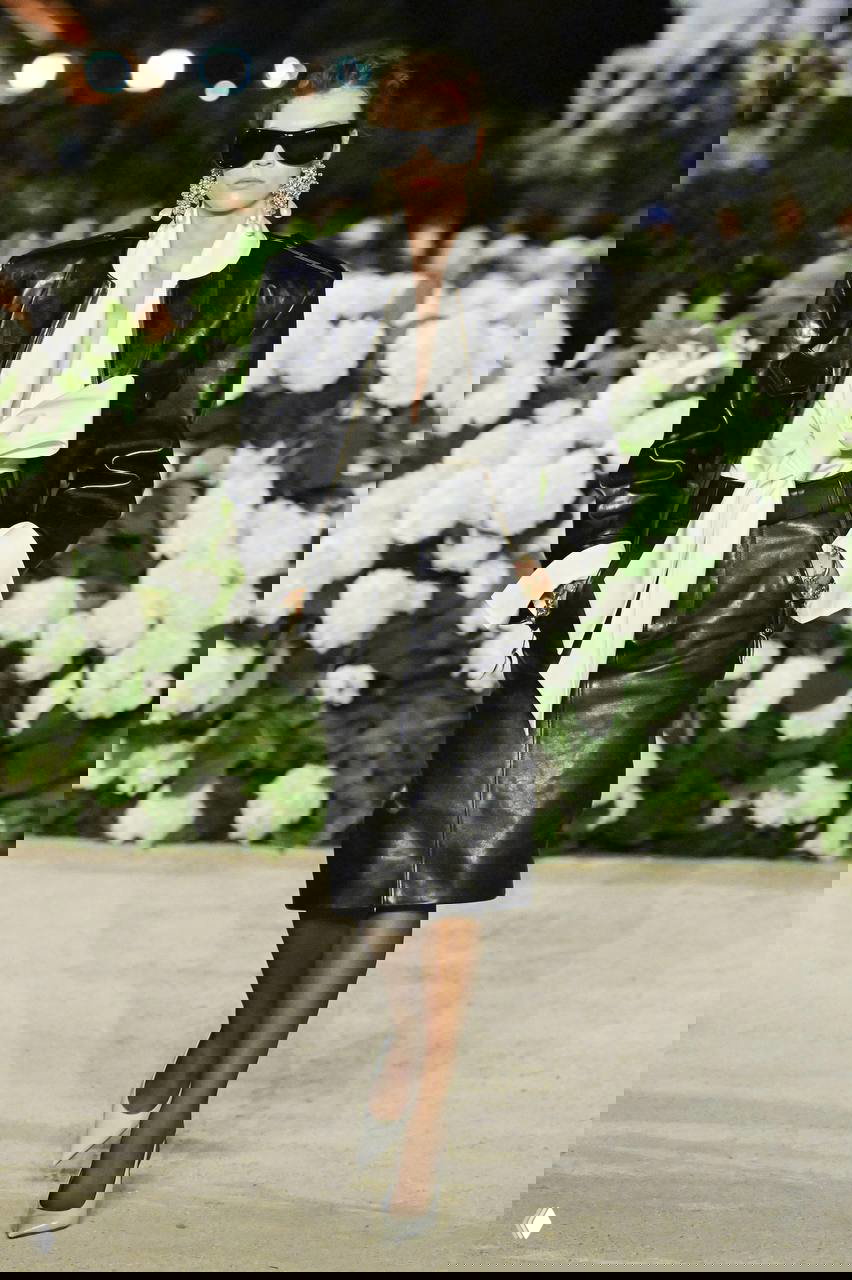 Модель в кожаном жакете с мощными плечами и строгой белой блузой на фоне цветов — сочетание дерзости и кутюрных деталей в тенденциях этого сезона.