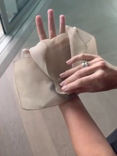 Крупный план рук с кольцами, раскрывающих ткань сумочки — внимание к деталям аксессуаров и маникюру на фоне деревянного пола и стекла.