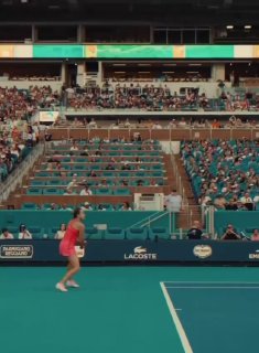 Кадр с корта во время матча на турнире в Майами: игрок в розовом платье выполняет передвижение по бирюзовому корту, трибуны частично заполнены.