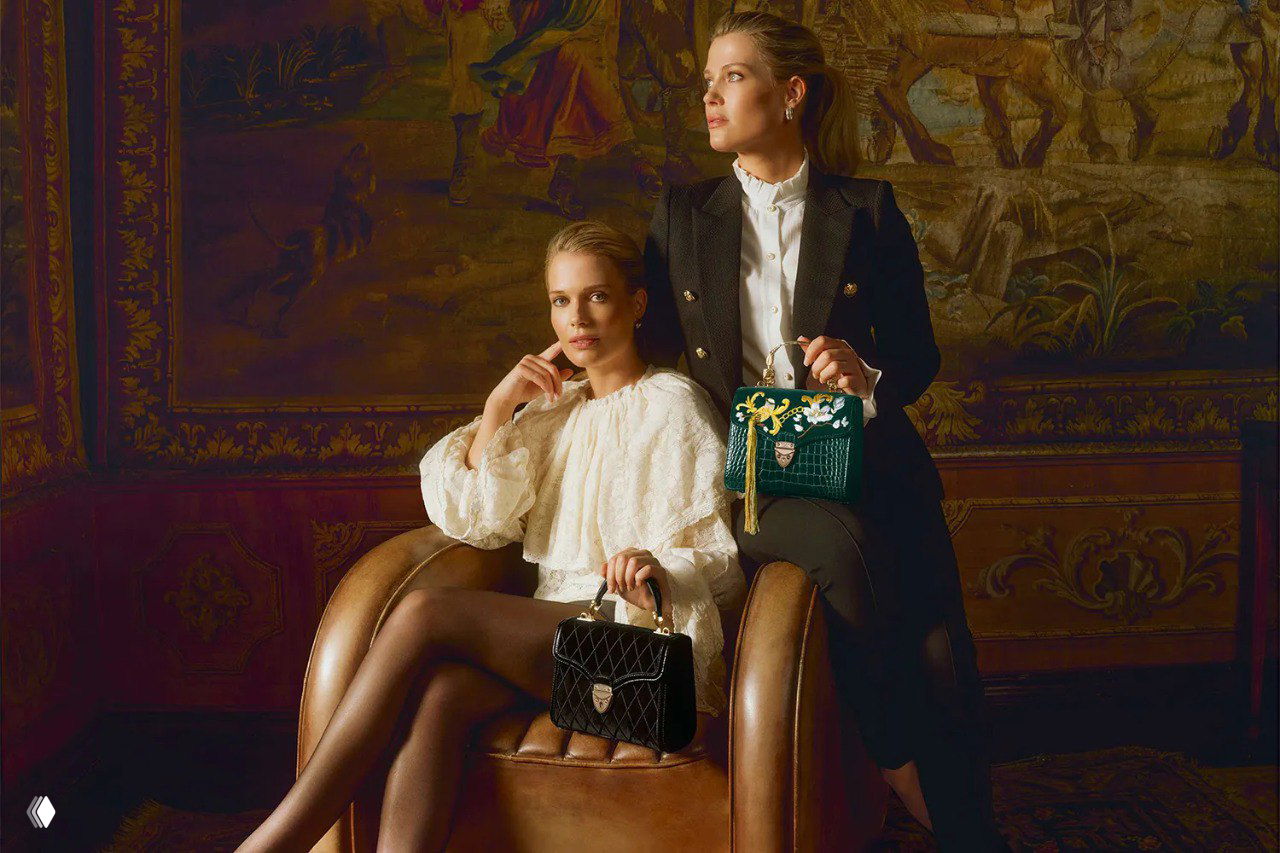 Элиза и Амелия Спенсер позируют в классическом зале с декоративной гобеленовой стеной, держа чёрную и зелёную сумки Aspinal.