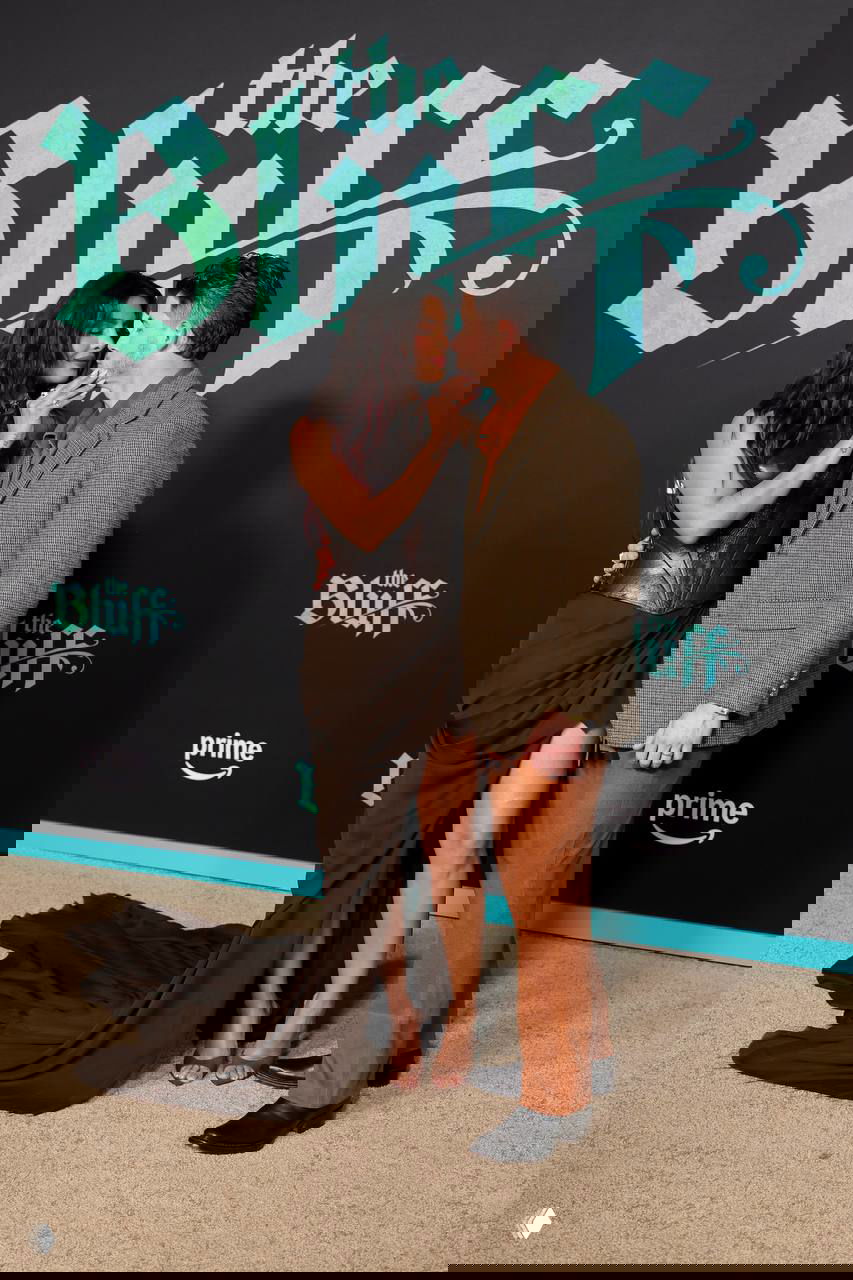 Приянка Чопра и Ник Джонас обнимаются на фоне логотипа «the Bluff»; платье с длинным шлейфом разложено за ними на красной дорожке.