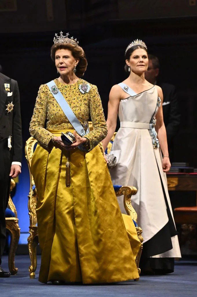 Две женщины в вечерних нарядах на сцене: одна в золотом платье сидит в кресле, другая в светлом платье стоит позади; формальная церемония с орденами и диадемами.