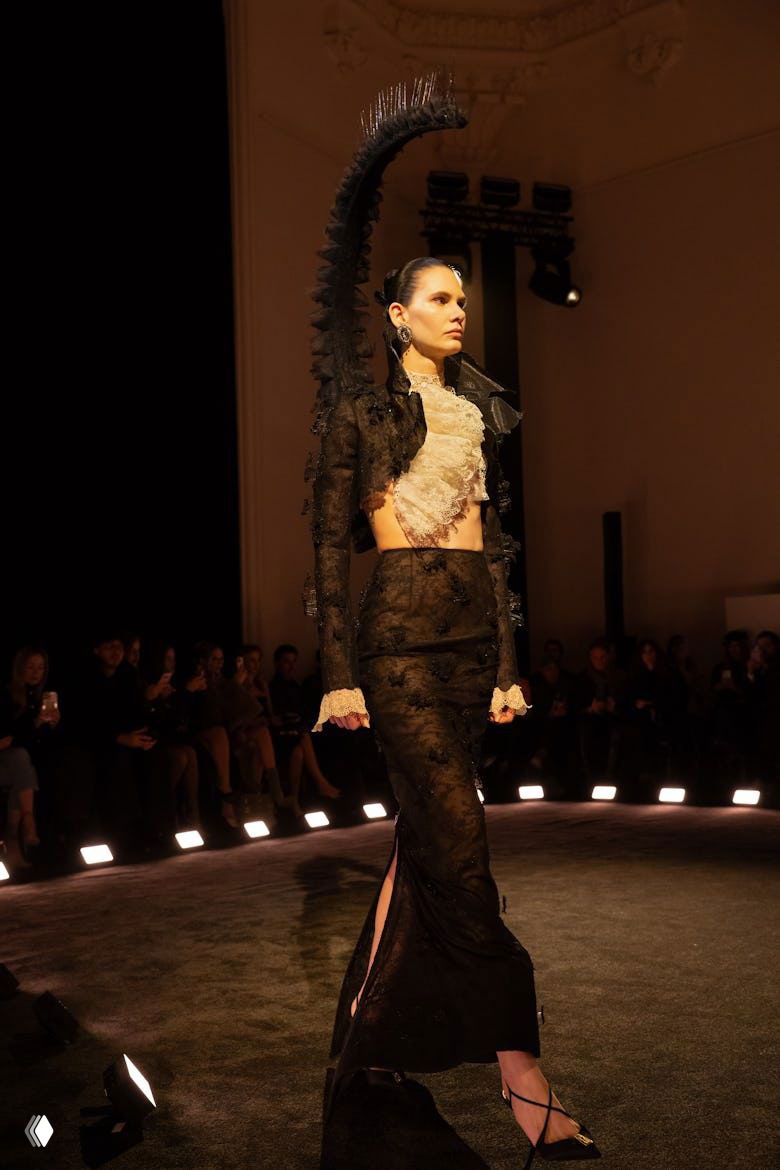 Модель в черном кружевном платье с высокой декоративной спинкой и скульптурным элементом, дефиле на круглом подиуме показа Schiaparelli.