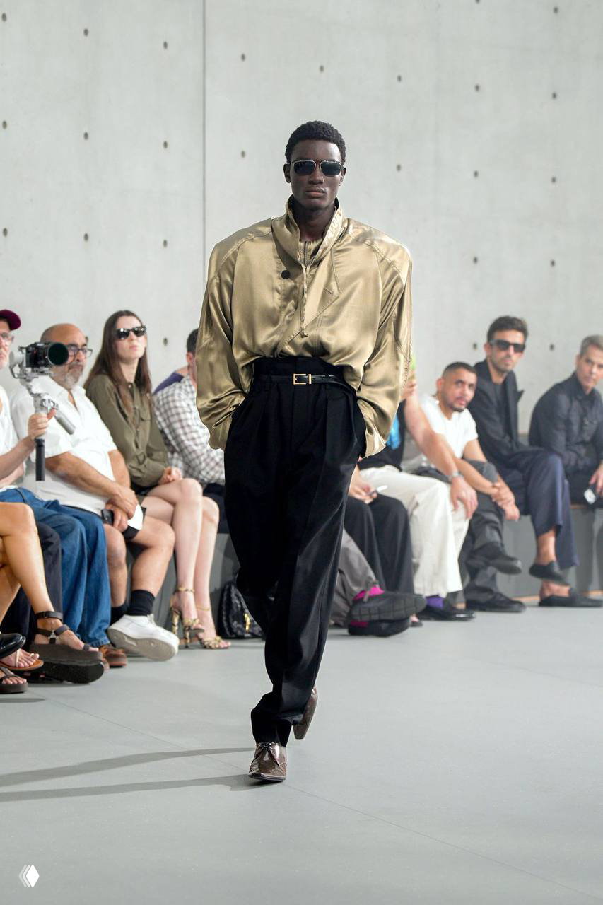 Модель в золотистом атласном пиджаке и чёрных брюках идёт по подиуму Saint Laurent; публика в первом ряду наблюдает показ и фотографирует образы.