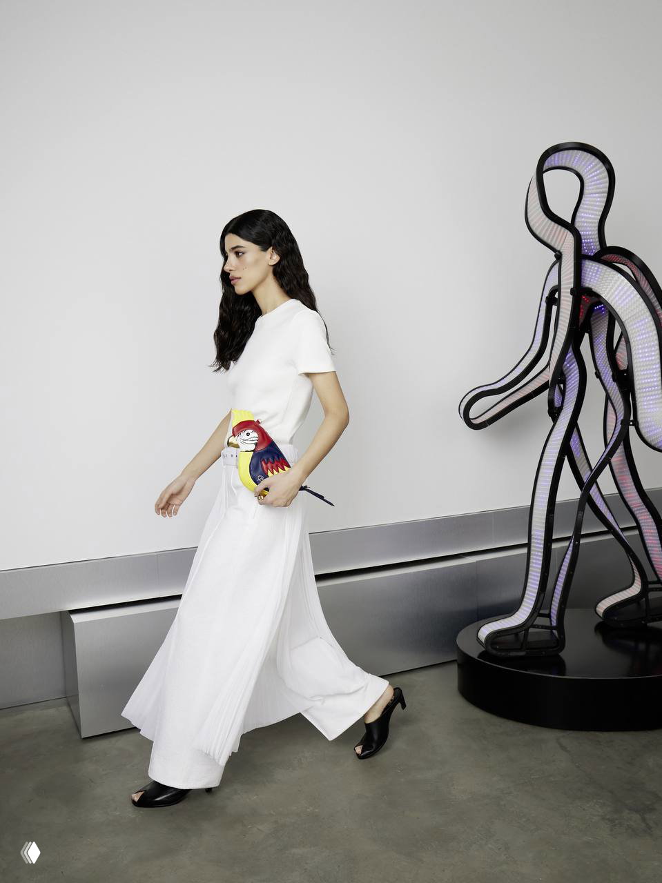 Модель в белом топе и широких брюках идет по студийному пространству мимо абстрактной скульптуры, кадр подчеркивает движение и аксессуар в руках.