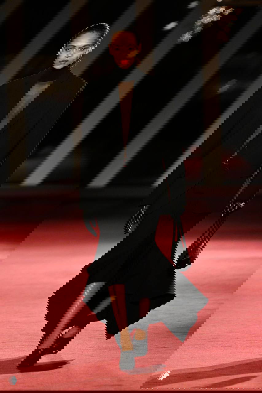 Модель в черном удлиненном пиджаке и струящейся юбке идет по подиуму на красном ковре — сочетание строгих линий и гламурного макияжа.