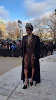 Показ: модель в прозрачном платье и накидке с украшенным головным убором идёт по дорожке, позади — зрители и городской пейзаж Парижа.