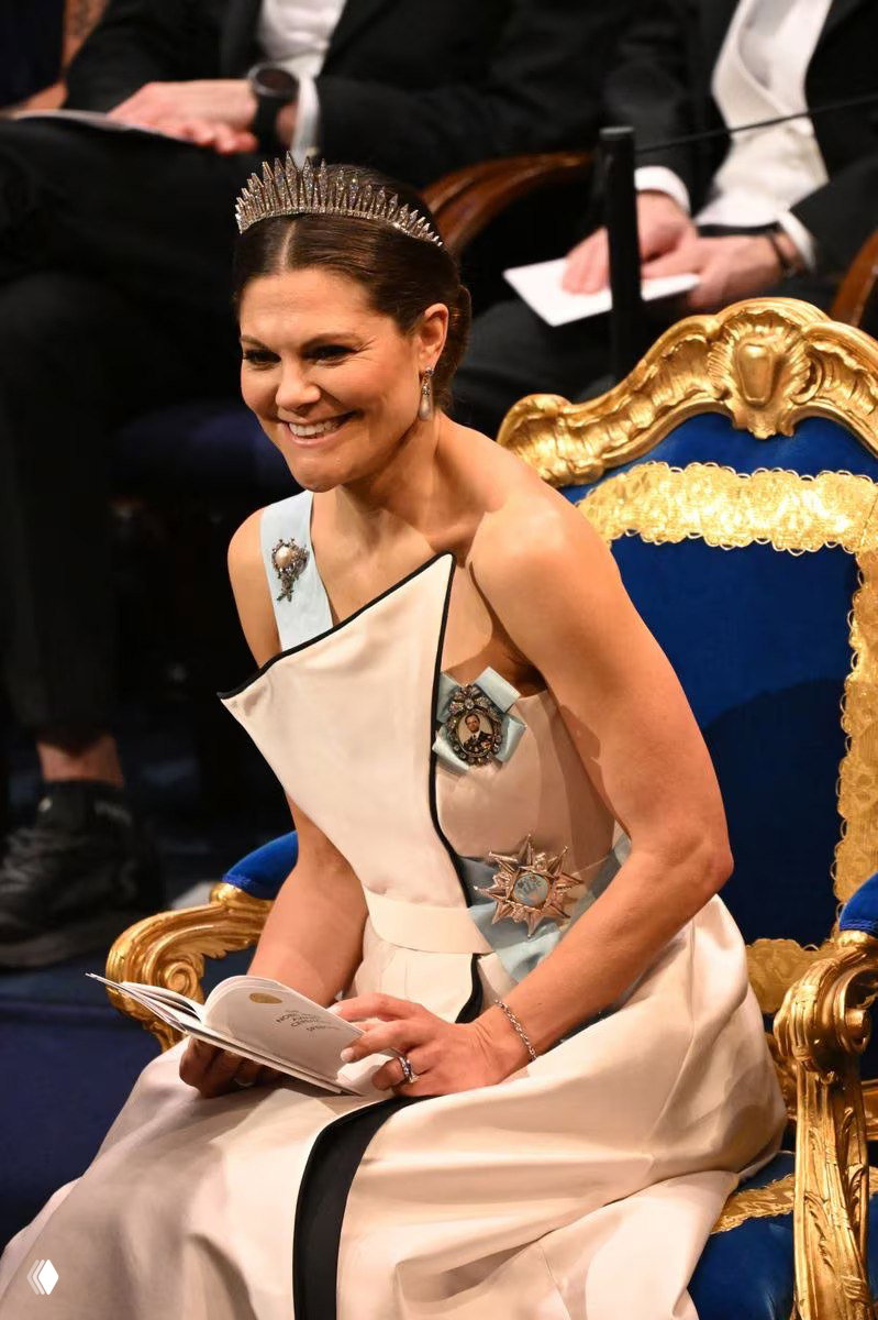 Крупный план женщины в светлом платье и диадеме: она улыбается во время церемонии, в руках держит программу, видны украшения и орден на плечевой ленте.