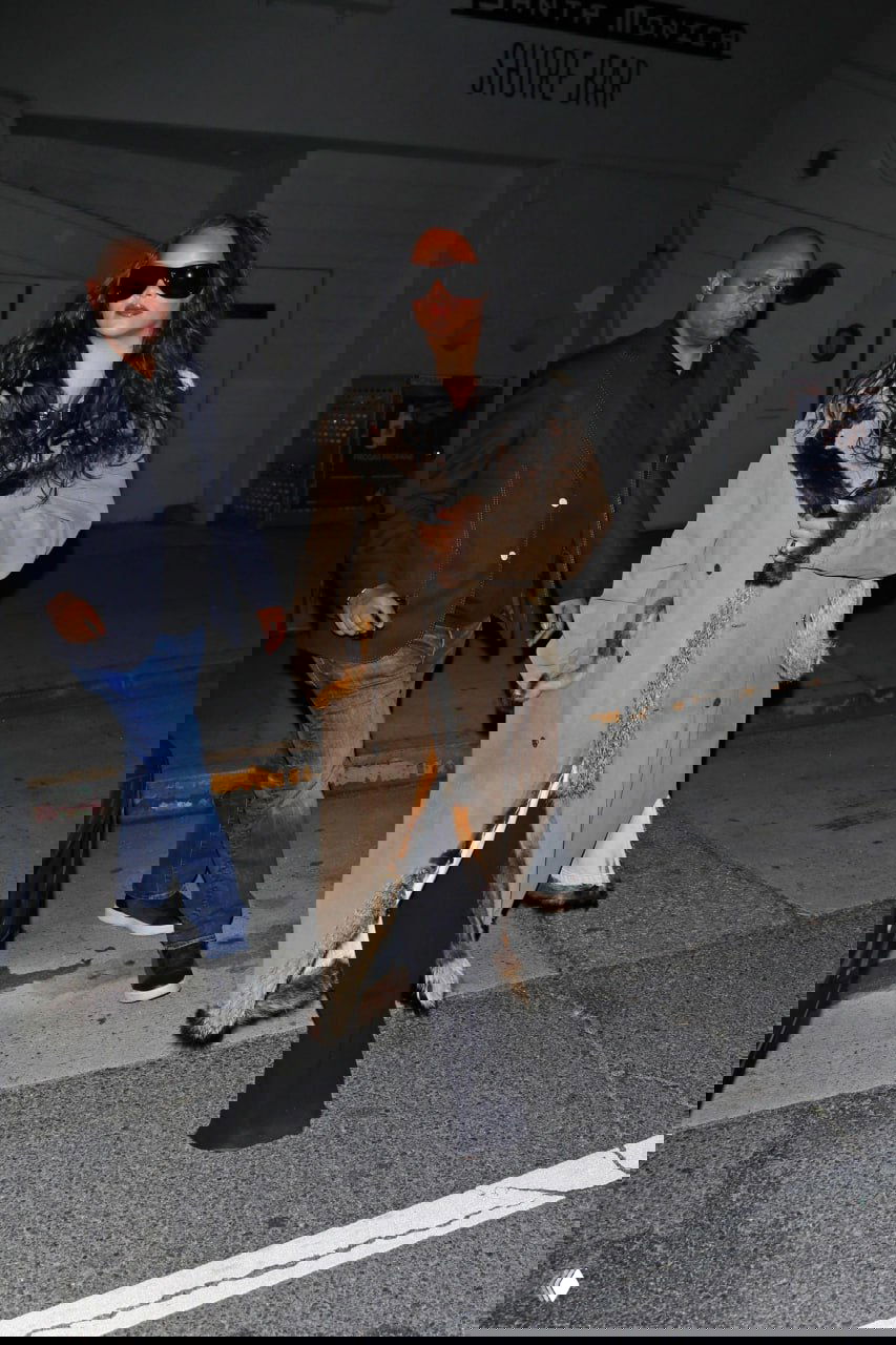 Ночная уличная фотография: женщина в длинном меховом или отделанном пальто идет по тротуару с охраной возле бара, сценический городской свет.