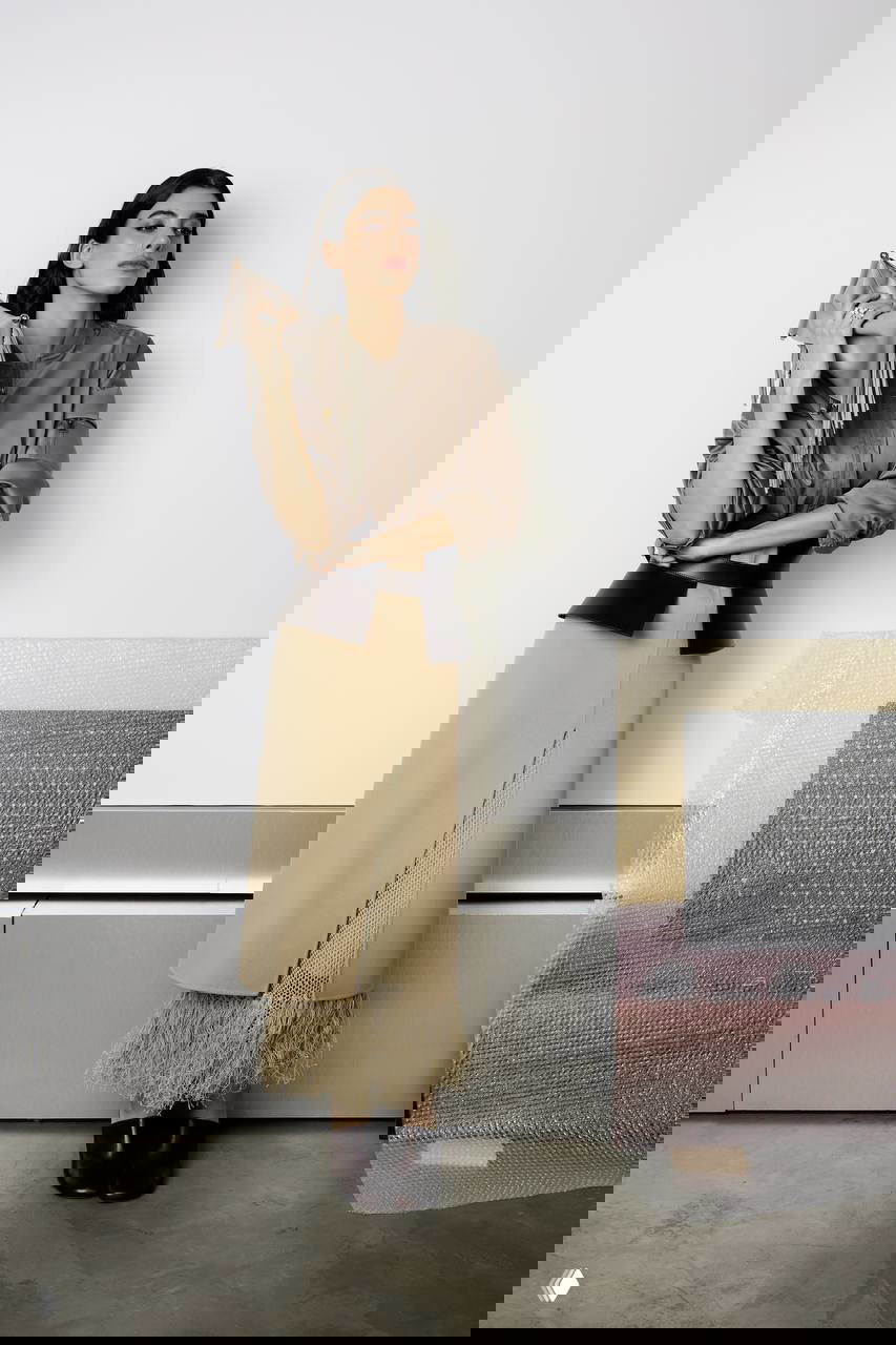 Модель в бежевой юбке с декоративной бахромой и коричневой рубашке позирует у стены, изображение демонстрирует сочетание фактур и деталей кроя.