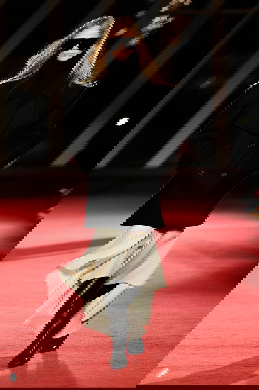 Модель в больших темных очках, черном пальто и белой юбке появляется на подиуме, демонстрируя сочетание практичности и роскошных деталей в коллекции.