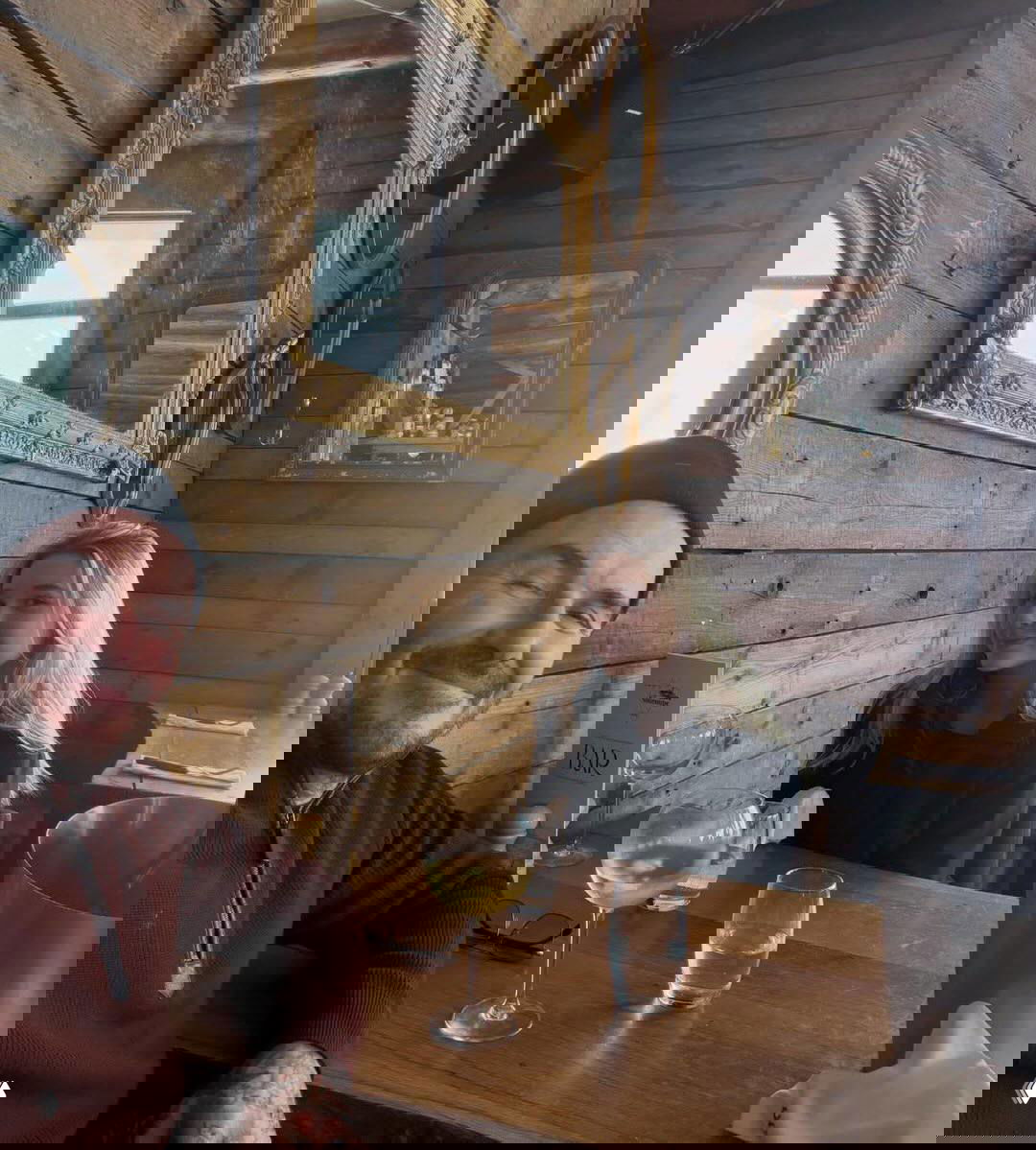 Мужчина и девушка за деревянным столом у стены, украшенной старинными зеркалами; бокал белого вина и альпийский антураж.