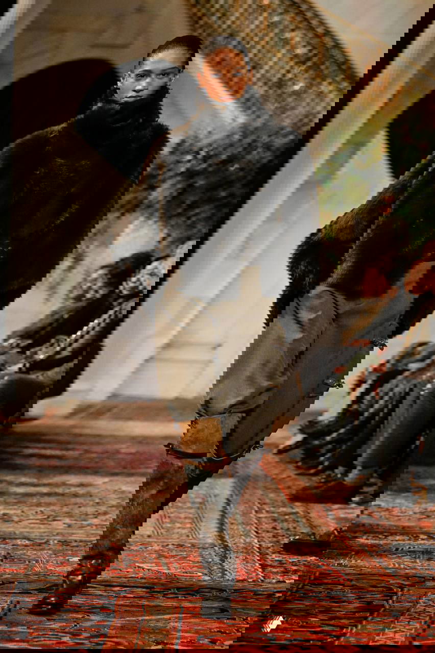 Модель в тёмной объёмной шубе и свободных брюках с массивным поясом и высокими сапогами, земляная палитра и грубая фактура на подиуме.