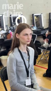 Модель сидит в гримерной зоне с бейджем на шее, вокруг видны стулья и зеркала — сцена подготовки к показу моды.