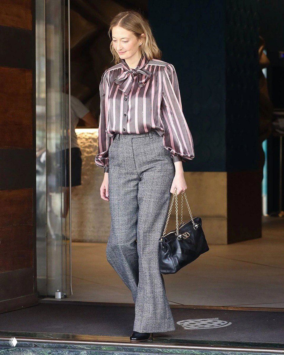 Женщина выходит из здания с чёрной сумкой Valentino Panthea на золотой цепочке; кадр показывает сочетание сумки с деловым или кэжуал-луком и удобной длиной ручки-цепи.