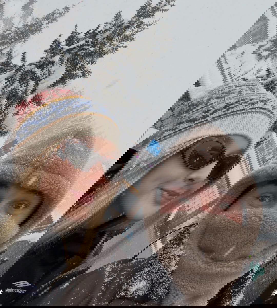 Девушка в вязаной шапке и мужчина в тёплой маске на фоне снежного пейзажа; крупный план улыбающихся лиц и падающий снег.