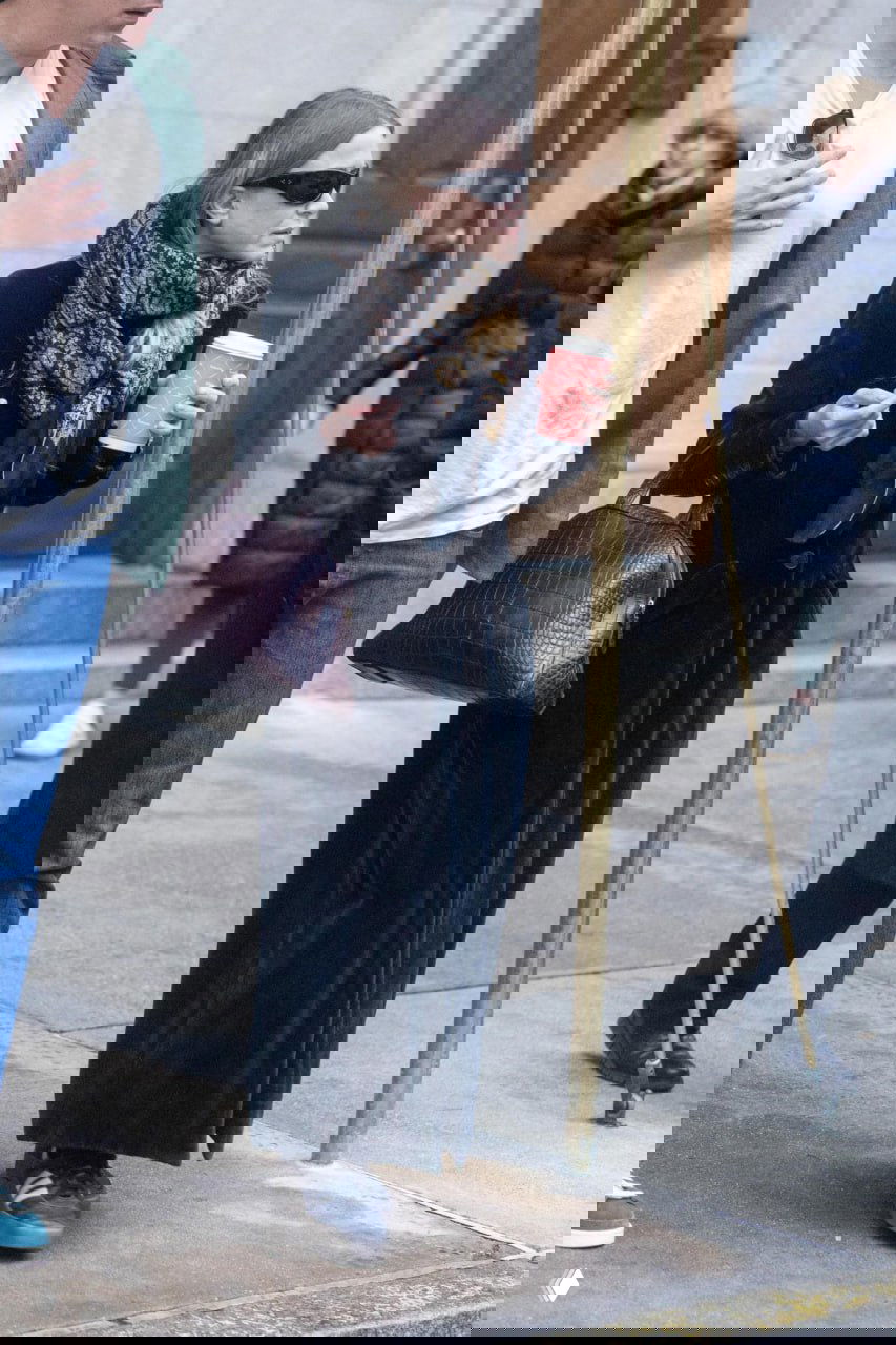 Городская сцена: женщина в длинном темном пальто, шарфе и солнечных очках держит стакан кофе, уличный стиль в прохладную погоду.