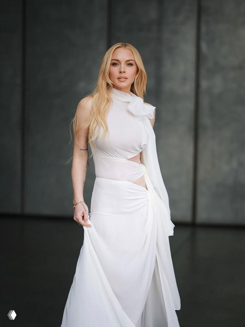 Крупный план Линдси Лохан в белоснежном платье с аккуратными вырезами и драпировкой, натуральный макияж и распущенные светлые волосы создают мягкий лук.