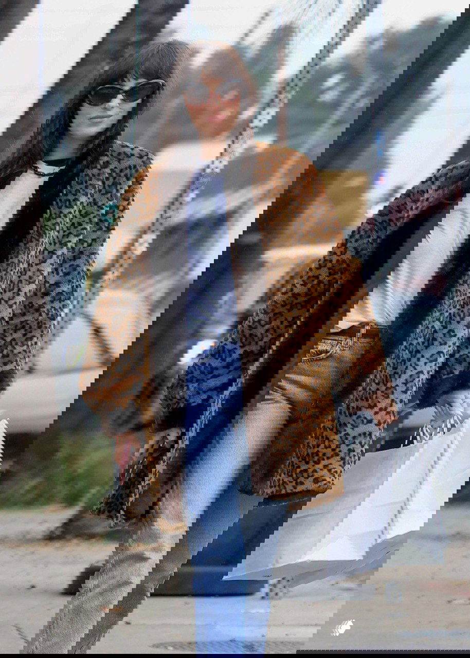 Крупный план в леопардовой шубе и солнцезащитных очках-«кошках», держит пакеты покупки на городском тротуаре.