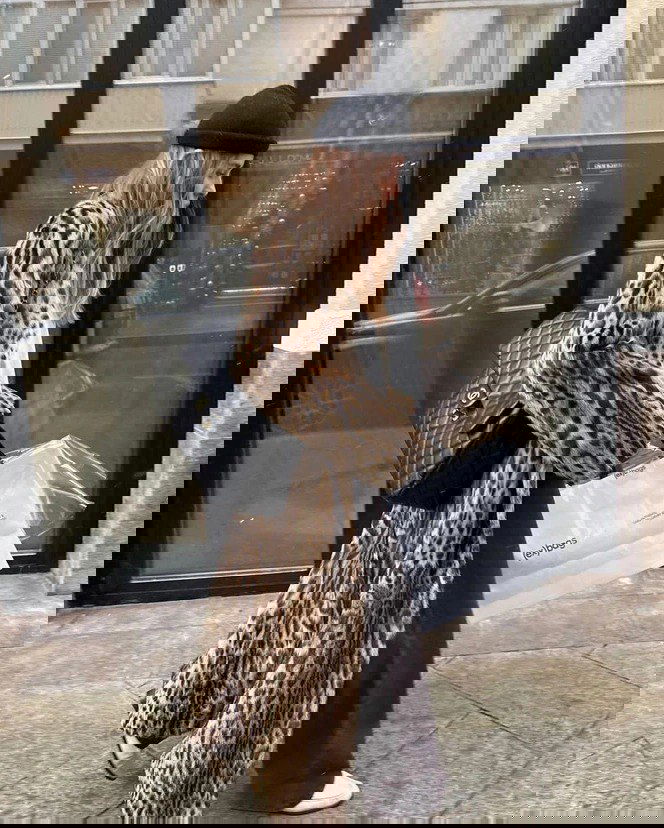 Длинное леопардовое пальто на прохожей в шапке, образ в анималистичном принте на фоне городского витрины и сумки Chanel