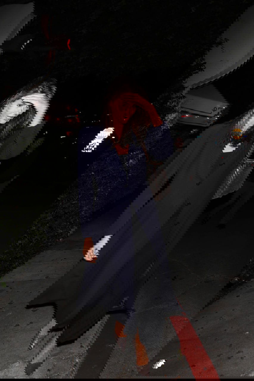 Женщина в длинном темном плаще идет по ночной улице, жест рукой к лбу от света камер, классический вечерний силуэт и прохожие.