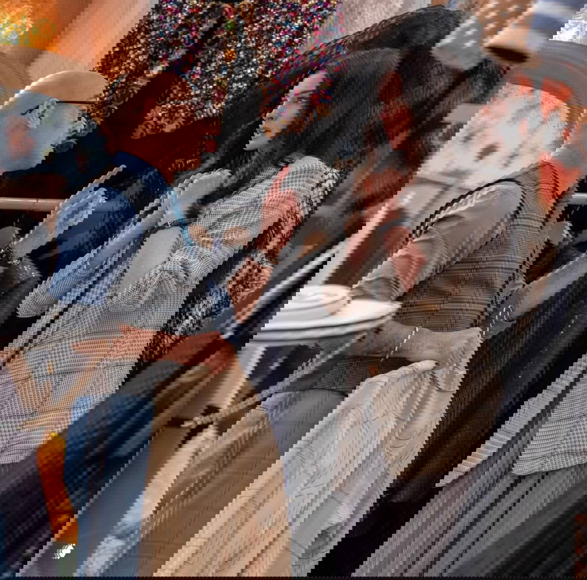 Съёмочный кадр в гардеробе: мужчина в жилете держит одежду и объясняет что‑то молодой женщине в пиджаке и длинной юбке.