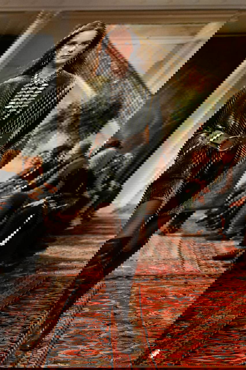 Модель в бархатном оливковом платье с крупными металлическими украшениями на груди, пояс и высокие сапоги — роскошный текстиль и металл.