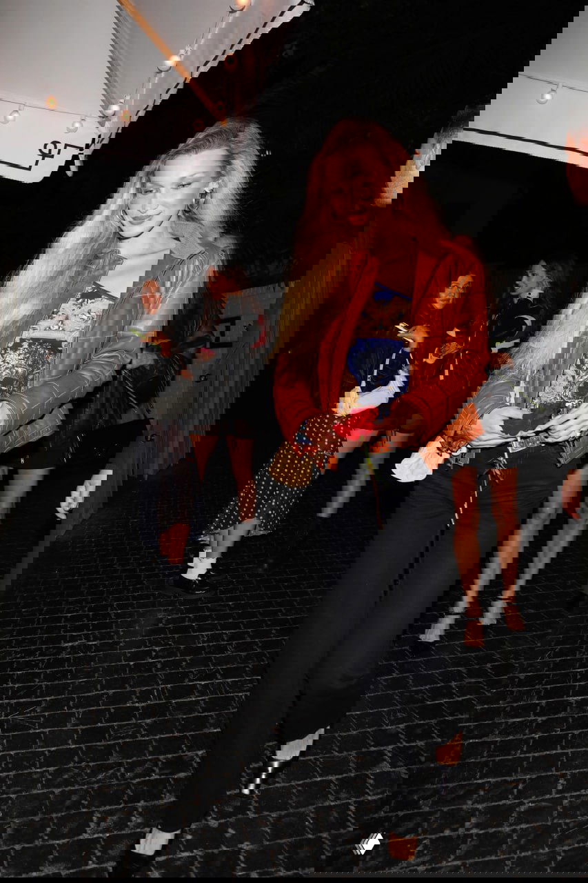 Женщина с длинными светлыми волосами в коричневой кожаной куртке и черных брюках идет по мокрой брусчатке, ночной уличный кадр.