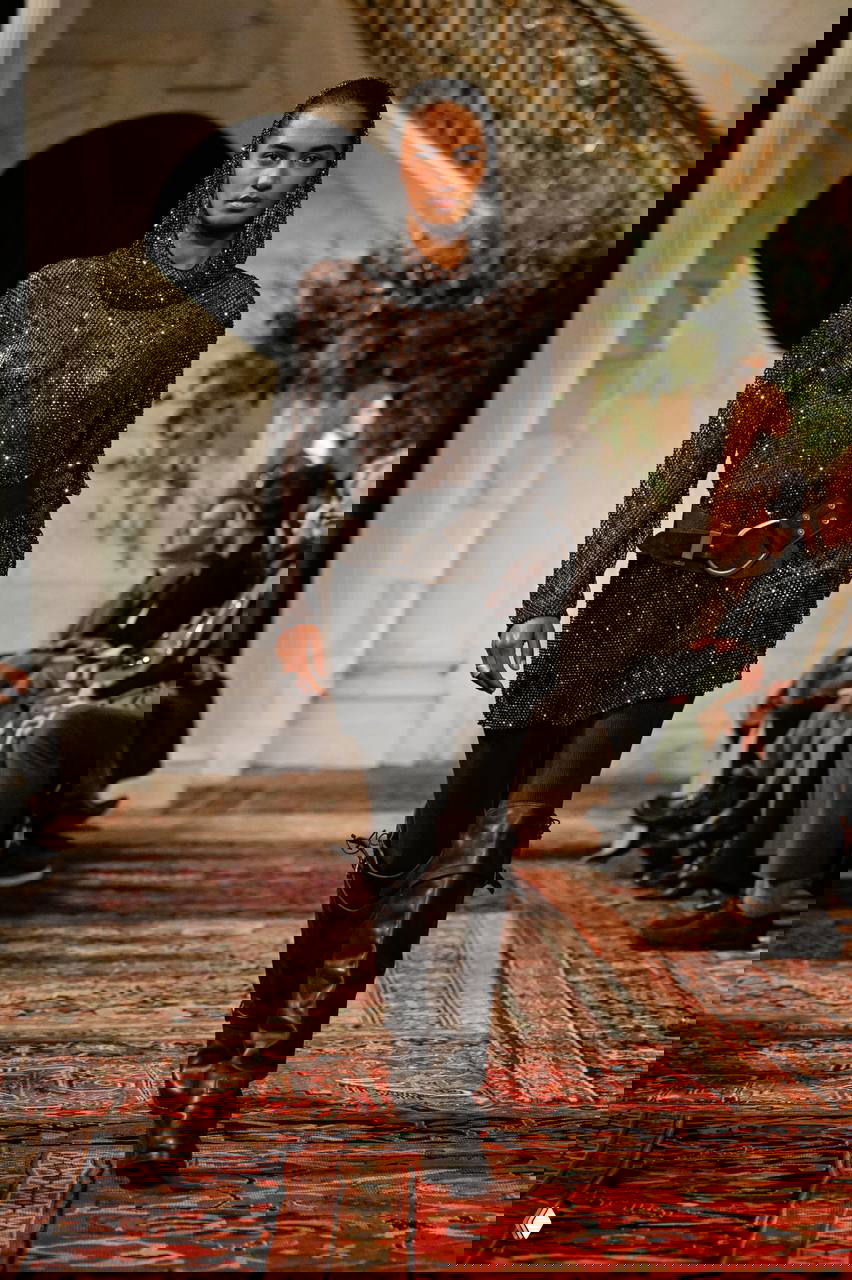 Модель в прозрачном блестящем худи со стразами, ремень на талии и сапоги, сочетание прозрачности, блеска и грубой обуви на подиуме.