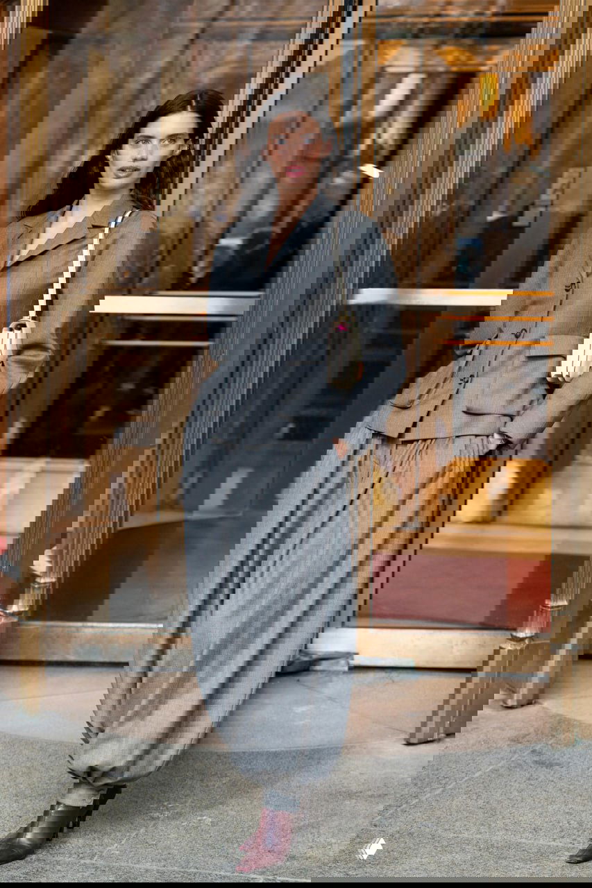 Модель в сдержанном сером костюме и широких шаровидных брюках на фоне дверей отеля, дневной городской стритстайл с классическими акцентами.