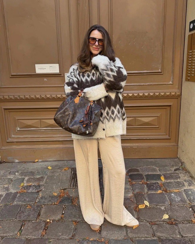 Стильный уличный образ: женщина в коричнево‑белом скандинавском свитере с крупным геометрическим узором и светлыми широкими брюками на брусчатке города.