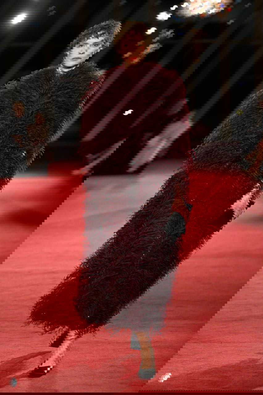 Модель в длинном бордовом пальто с выразительной перьевой фактурой выходит на подиум, подчеркивая игру объема и текстуры в коллекции.