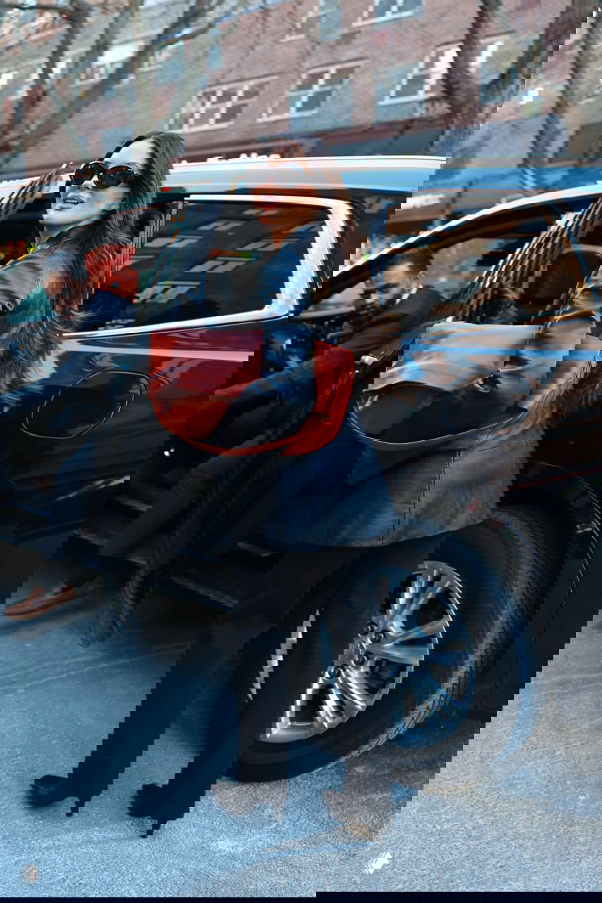 Женщина в черном кожаном пиджаке, солнцезащитных очках и с крупной коричневой сумкой садится в автомобиль — снимок в городском окружении.