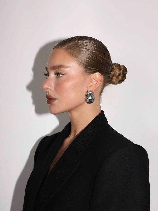 Профиль женщины в низком пучке с открытыми ушами, линия шеи и скулы хорошо видны; минималистичный образ с серьгой на одно ухо и строгим чёрным жакетом.
