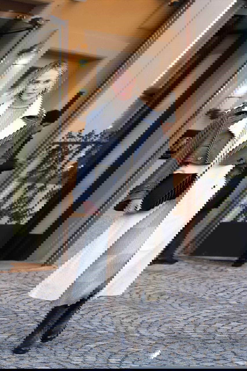 Дневной кадр: женщина в светлом пальто с кожаной вставкой и высокими ботинками, шаг по брусчатке у входа в здание — минималистичный образ.