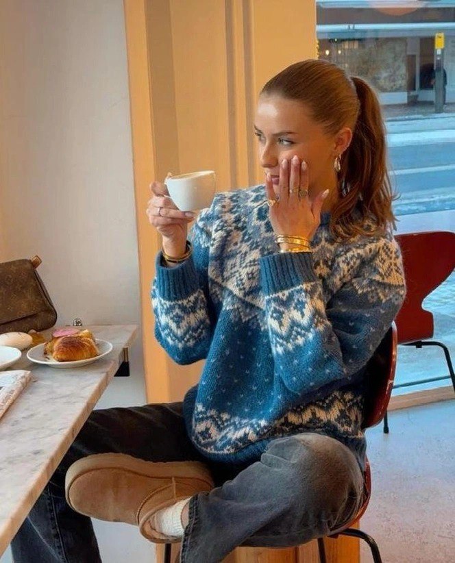 Женщина в голубом скандинавском свитере с белым орнаментом сидит в кафе с чашкой, сочетая уютный зимний стиль и повседневную элегантность.