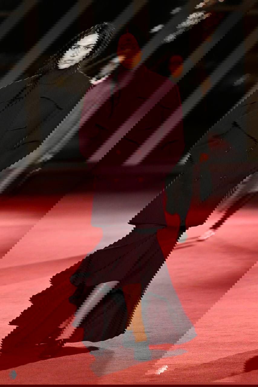 Модель в темном пальто и волнистой длинной юбке шагает по красной дорожке показа Michael Kors, сочетая строгие силуэты и драпировки.