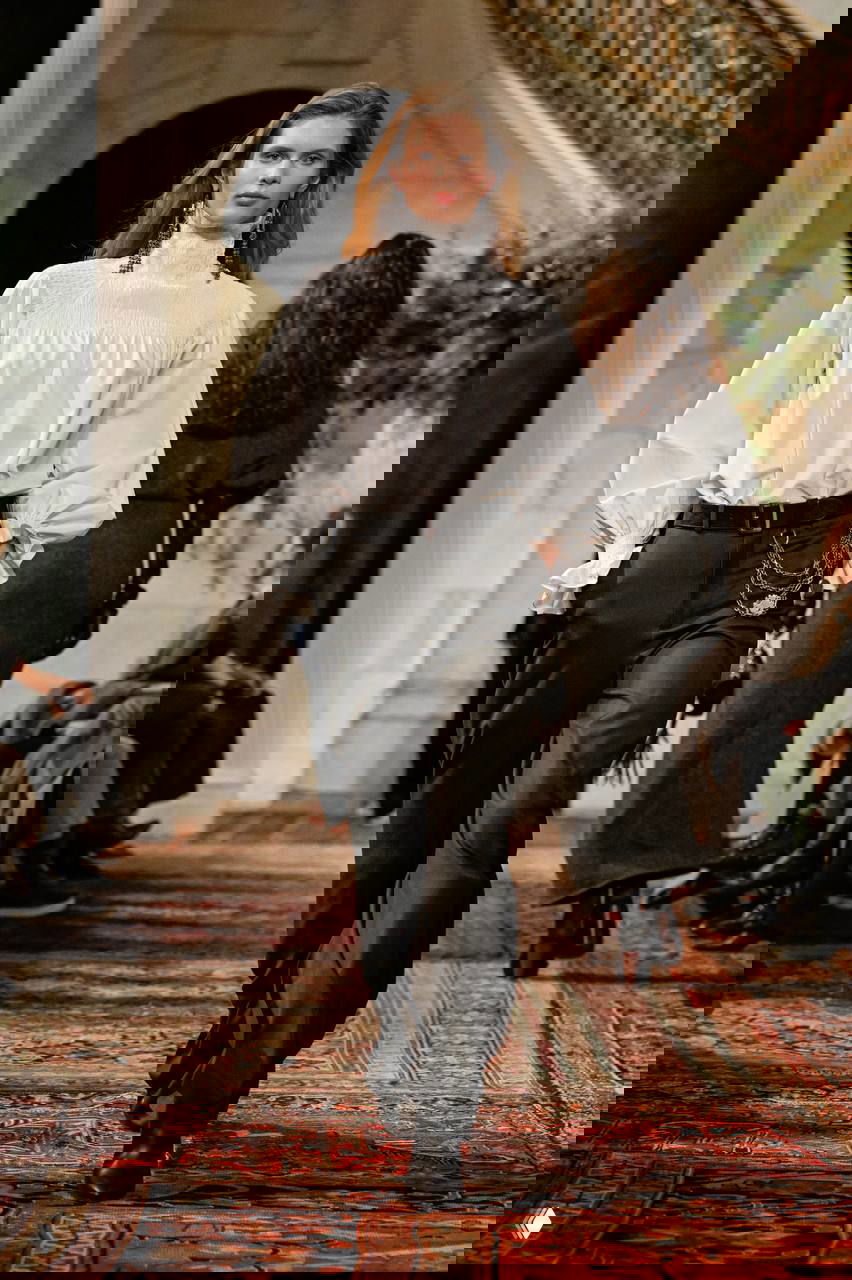 Модель в белой блузе с высокой горловиной и чёрных брюках, цепочки на поясе и классический силуэт в благородной земляной палитре коллекции.