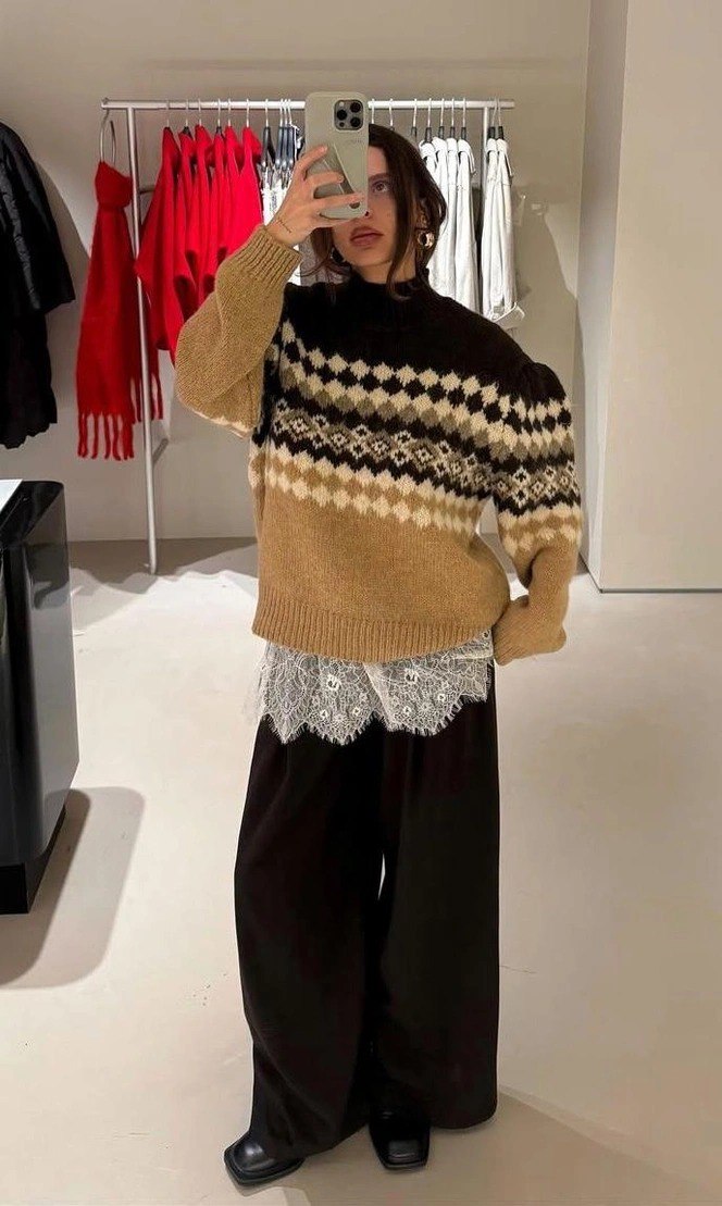Фотоснимок в примерочной: девушка в коричнево‑бежевом свитере с контрастным орнаментом и кружевной юбкой, демонстрирующая сочетание фактур и слоёв.