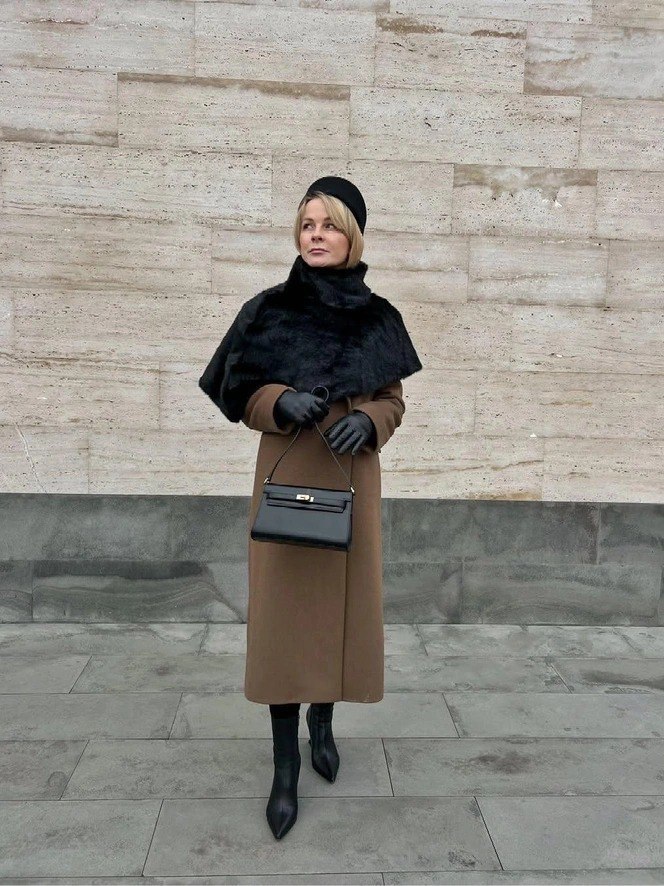 Женщина в коричневом длинном пальто и тёмном укороченном кейпе на фоне бежевой каменной стены; образ с сумкой и кожаными перчатками для улицы.