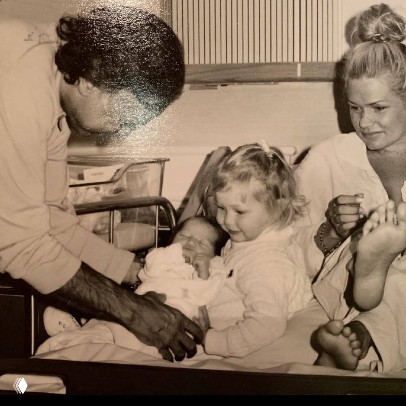 Чёрно-белое архивное фото: взрослая женщина, малыш в родильном отделении и маленькая девочка, наблюдающая за младенцем; трогательный семейный момент.