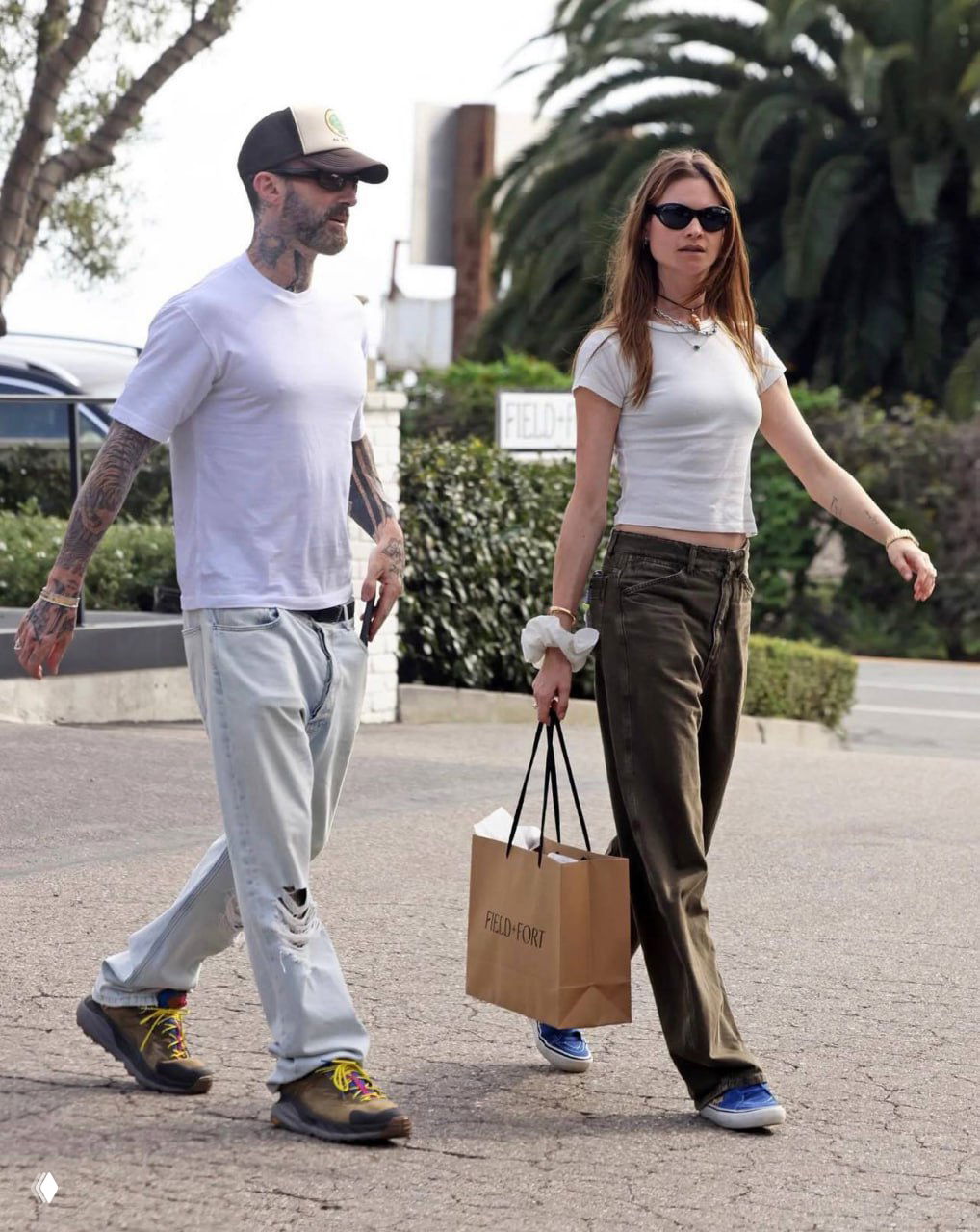 Пара идет по улице: женщина в свободных брюках и белой футболке, мужчина в светлой футболке и бейсболке; повседневный уличный лук.