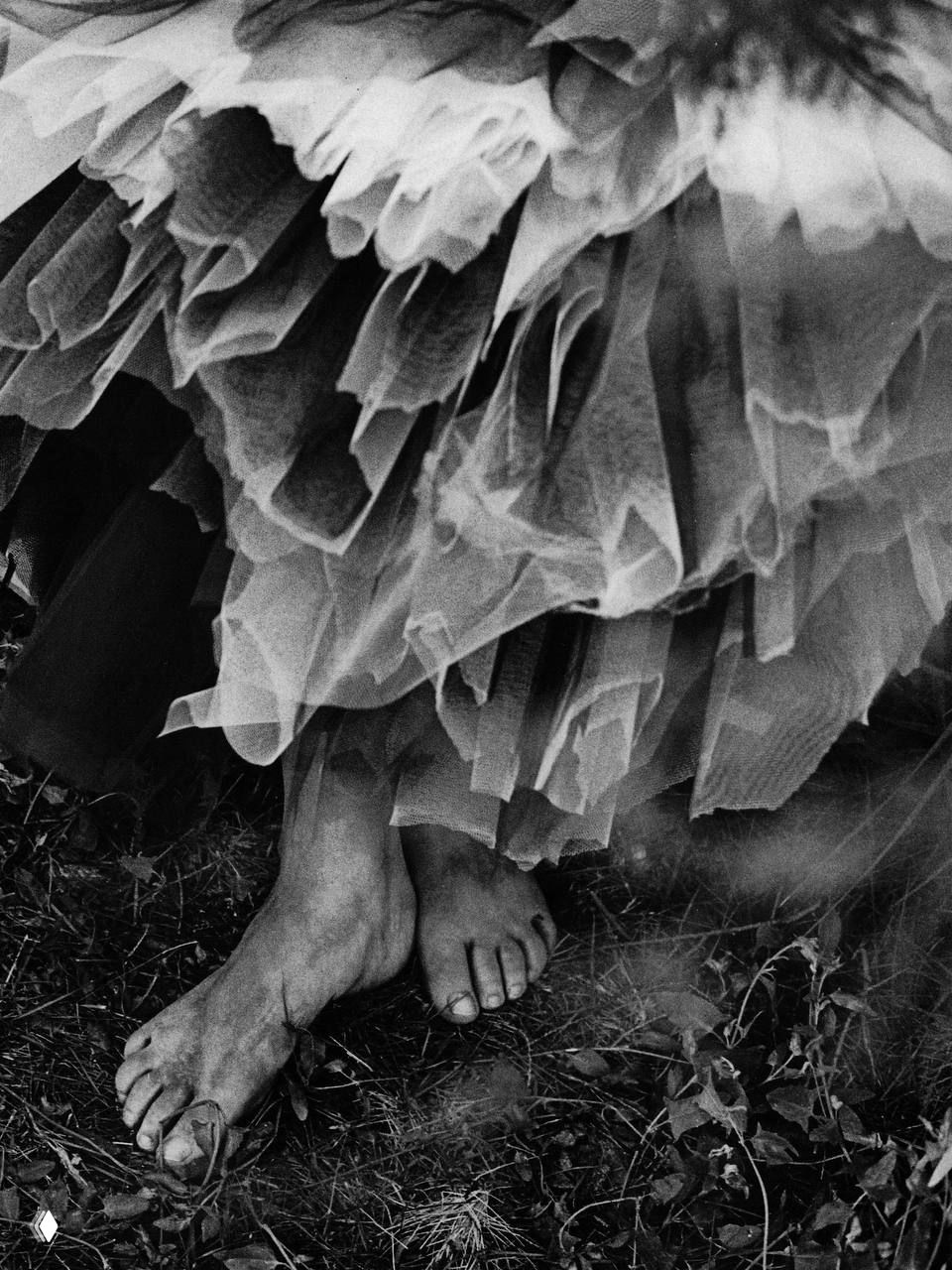 Крупный план: босые ноги модели под многослойной фатиновой юбкой, чёрно‑белая фактура ткани и текстуры земли в винтажном кадре.