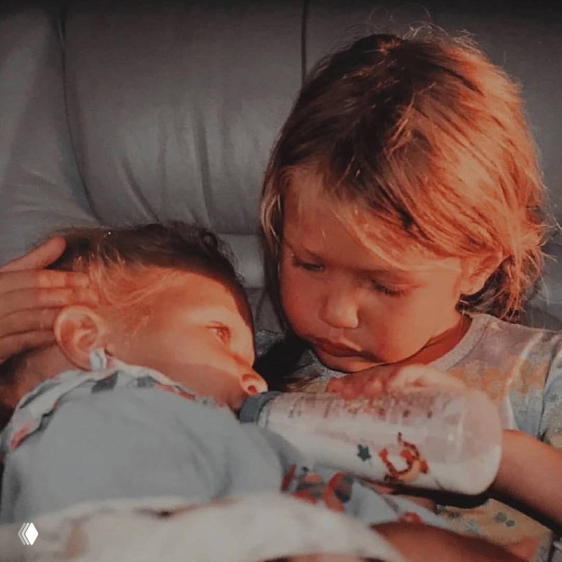 Ретро-снимок двух маленьких детей: одна девочка держит бутылочку для кормления, другая внимательно помогает; тёплые детские воспоминания на фото.