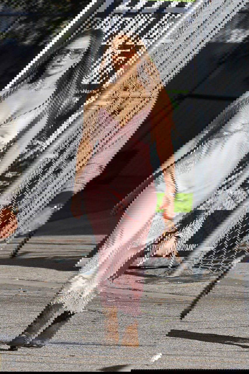 Женщина в розовом сатиновом платье и ковбойских ботинках идет по солнечной улице, контраст романтичного платья и грубой обуви.