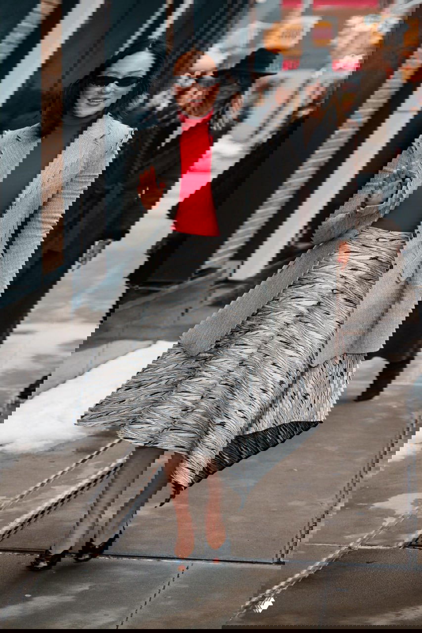 Женщина в сером пиджаке и пышной асимметричной юбке идёт по городской улице в солнцезащитных очках и красном свитере. Фотография уличного папарацци.