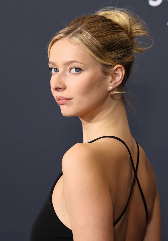 Крупный план женщины на премьере с пучком, минимальным макияжем и перекрёстными бретелями чёрного платья