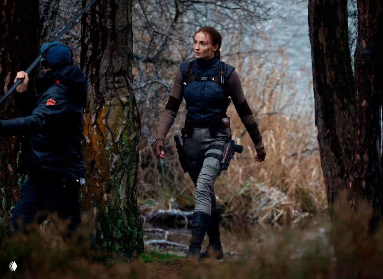 Актриса в костюме Лары Крофт идёт по лесной кромке у воды, рядом съёмочная группа и оборудование на фоне.