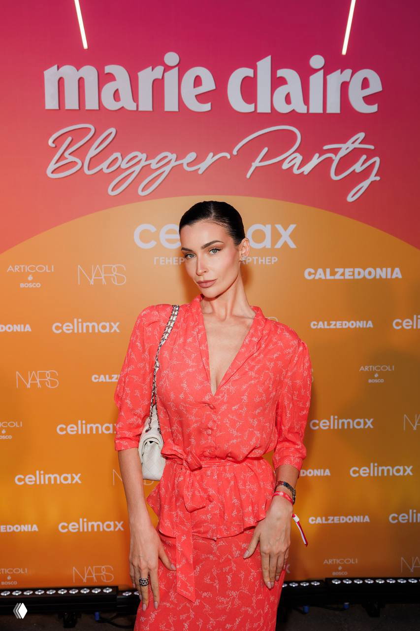 Женщина в коралловом платье позирует у брендовой фотозоны Marie Claire Blogger Party; видно оформление и часть световой инсталляции.